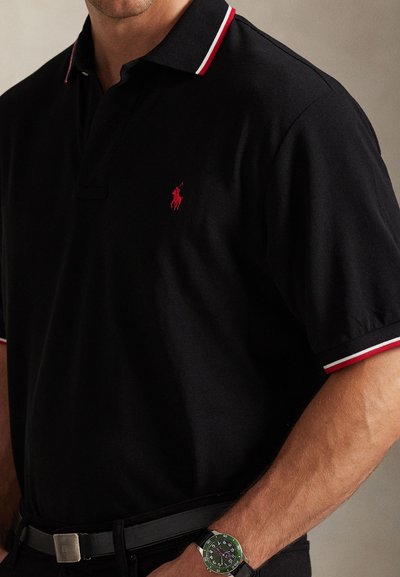 Férfi fekete pólóingben, piros-fehér gallérral és ujjcsíkokkal, fekete övvel és zöld keretes karórával.