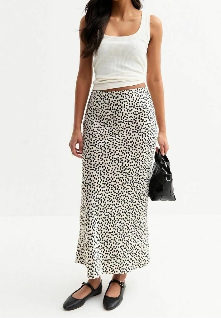 Femme portant un débardeur blanc et une longue jupe blanche à pois noirs, tenant un petit sac à main noir et portant des ballerines noires.