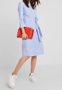 Niebieska sukienka w paski z paskiem w talii, połączona z fakturowaną czerwoną kopertówką i białymi trampkami. Sukienka ma zapięcie na guziki i rękawy trzy czwarte.
