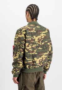 Camo bomber jacket in green, brown, and beige. Has a ribbed collar and cuffs, with a pocket on the sleeve featuring a red tag. Textured fabric.