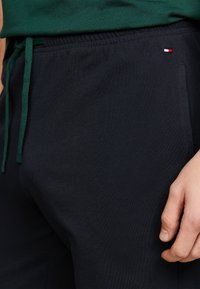 Pantaloni jogger blu navy realizzati in materiale morbido, con vita elasticizzata e stringa verde. Piccolo logo decorativo in rosso, bianco e blu vicino alla tasca.