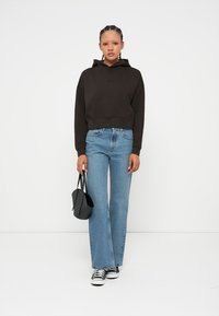 Fiatal nő fekete, rövidített kapucnis pulóverben, kék, laza fazonú farmert visel, fekete tornacipőt hord, és egy kis fekete táskát tart, miközben egy fehér háttér előtt áll.