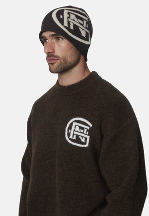 Hombre que lleva un suéter marrón oscuro y un gorro negro, ambos con un gran logotipo circular blanco con letras estilizadas.
