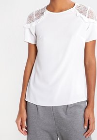 Haut blanc à manches courtes avec des détails en dentelle aux épaules, col rond, texture lisse et coupe décontractée. Associé à un bas gris.