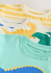 Kolm t-särki on kuhjatud. Ülemine: kollane valgete triipudega. Keskmine: kreemjas dinosauruse mustriga. Alumine: teal siniste ja kollaste aktsentidega.