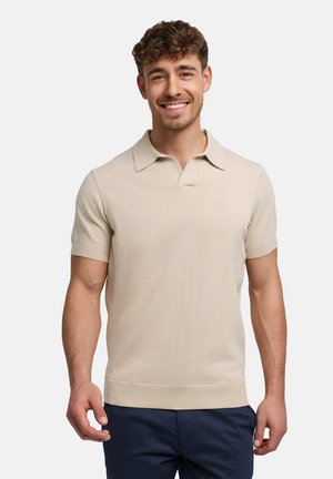 Glunderende man met kort krullend haar, gekleed in een beige gebreide polo met korte mouwen en donkerblauwe broek, staand tegen een eenvoudige witte achtergrond.
