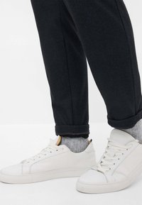 Witte leren sneakers met vlakke zolen, gecombineerd met grijze geribde sokken. De broek is donker met een subtiele textuur en is op de enkels omgeslagen.