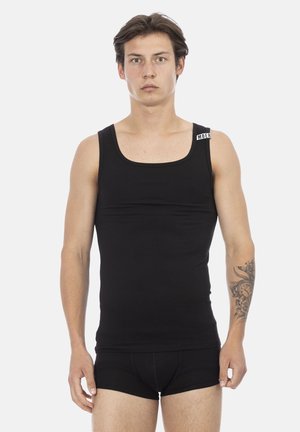 Jonge man met kort bruin haar, gekleed in een zwart tanktop en zwarte boxershorts, staand tegen een effen witte achtergrond.