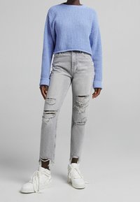 Personne portant un pull en tricot bleu clair, un jean gris clair taille haute déchiré, et des baskets montantes blanches se tenant sur un fond gris.