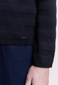 Pull en maille à rayures bleu marine avec des lignes horizontales texturées. Comprend une petite étiquette de marque métallique près de l'ourlet. Détail de poignets côtelés et doux.