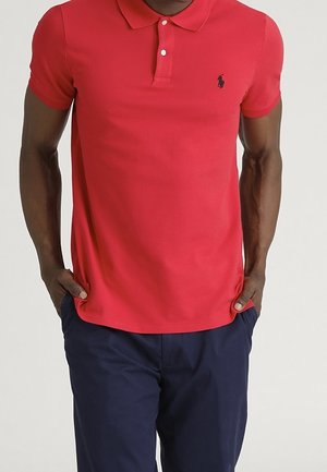 Homme portant un polo rouge à manches courtes avec un petit logo noir de cheval et un pantalon bleu marine, les mains dans les poches.