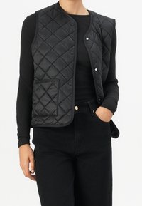 Osoba v čern�é prošívané bezrukávé vestě přes černý dlouhý rukáv, s černými kalhotami a jednou rukou v kapse.