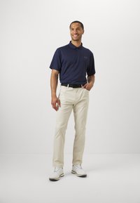 Cămașă polo albastră închis cu un model texturat, asortată cu pantaloni bej deschis și pantofi albi. Include un cureaua neagră și mâneci scurte.