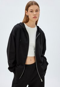 Sudadera con cremallera negra hecha de un tejido suave, con un corte holgado, capucha con cordón y bolsillos laterales, combinada con una camiseta corta blanca.