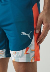 Pantalones cortos deportivos en color teal con acentos laterales en naranja. Presenta un patrón geométrico en el dobladillo y un logo reflectante de Puma en el muslo.