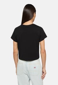 Schwarzes, kurzes T-Shirt mit kurzen Ärmeln, aus Baumwolle. Sichtbare Nähte um den Halsausschnitt und die Schulternähte, kombiniert mit hellblauen Jeans.