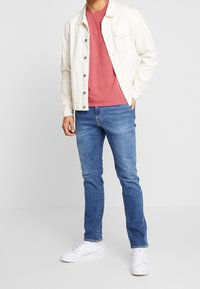 Mann in einer weißen, knöpfbaren Jacke über einem roten T-Shirt, blauen Jeans und weißen Sneakers, der mit einer Hand in der Tasche steht.