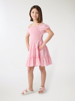 Ragazza giovane in piedi con le mani sui fianchi che indossa un vestito rosa scollato e sandali bianchi con fibbie rosa.