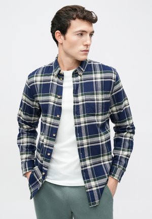Navy blue and green plaid flannel shirt with button-down collar, featuring light gray stripes and visible buttons, worn over a white shirt.