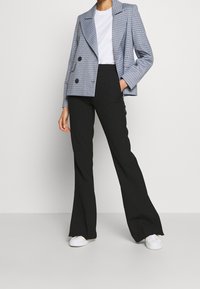 Grårutigt dubbelknäppt blazer med bred slag, parat med figursydda svarta byxor och vita sneakers, visad mot en neutral bakgrund.