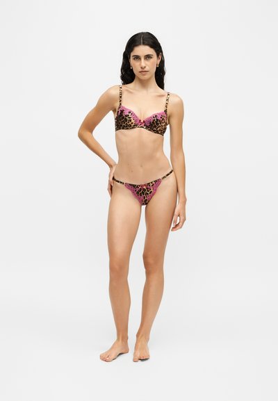 Femme aux longs cheveux foncés portant un soutien-gorge imprimé léopard et un string assorti avec des accents en dentelle rose, debout devant un fond uni clair.