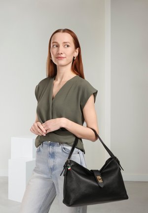 Sac à main en cuir noir avec une surface texturée, doté d'une fermeture à bouton dorée et d'une étiquette à rayures en guise d'accent. Le modèle porte un haut vert olive et un denim clair.