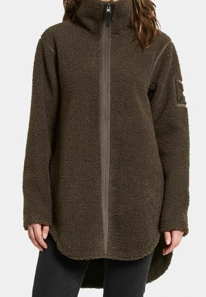 Persoon met een bruine fleecejas met hoge kraag, volledige rits aan de voorkant en lange mouwen, gecombineerd met een zwarte broek.