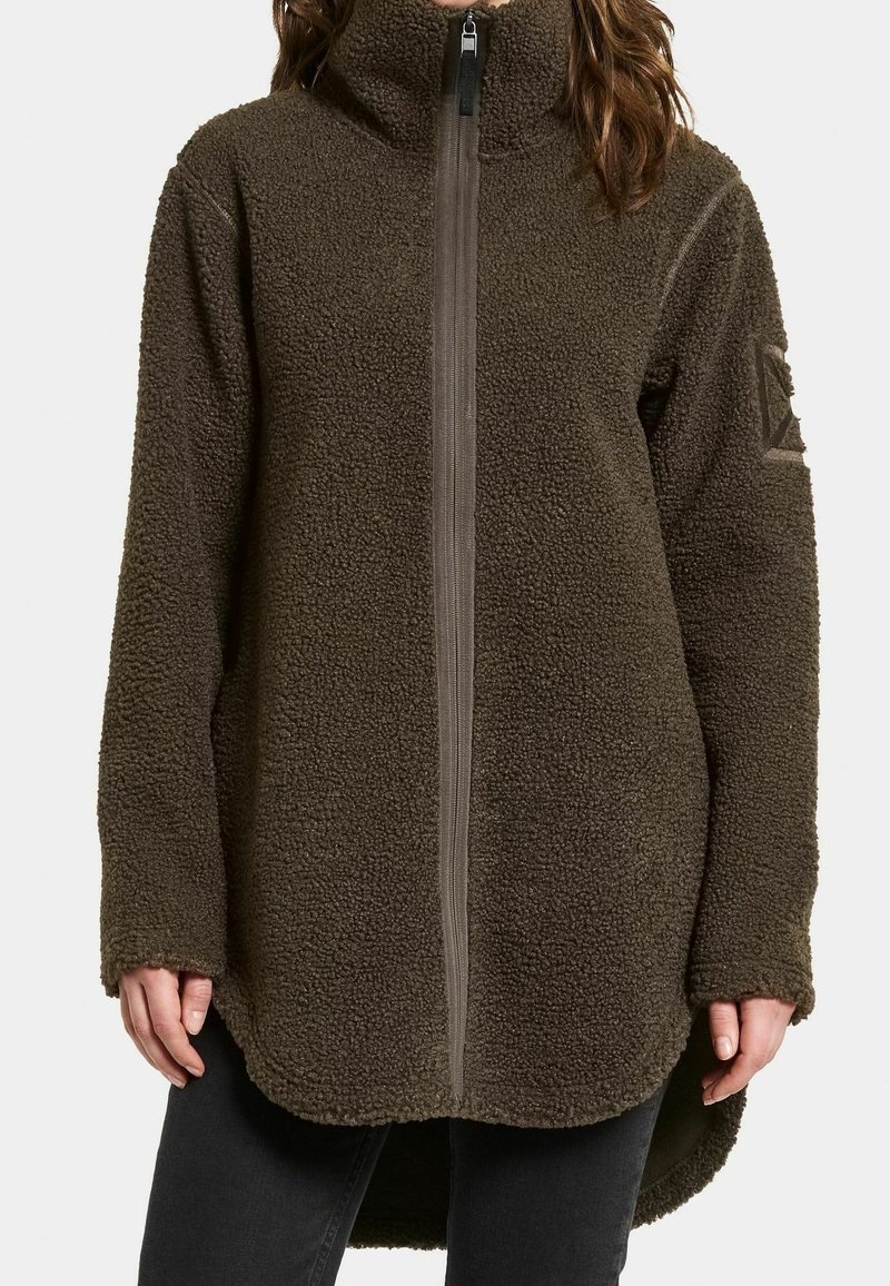 Personne portant une veste polaire marron à col montant, avec une fermeture éclair complète à l'avant et des manches longues, associée à un pantalon noir.