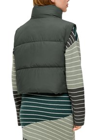 Dunkelgrüne Steppweste mit hohem Kragen, tailliertem Schnitt und glatter Textur; getragen über einem mehrfarbigen gestreiften Strickkleid.