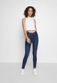 Prilagođeni bijeli crop top i tamnoplave uske traperice, izrađene od denima, uparene s bijelim tenisicama. Jednostavan dizajn bez vidljivih uzoraka.