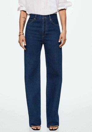 Vrouw draagt high-waist, wijde donkerblauwe denim jeans met een wit shirt en zwarte open-toe sandalen, staand tegen een effen achtergrond.