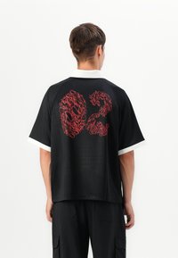 Black short-sleeve polo shirt with a white collar and red flame-patterned graphic on the back. Fabric appears smooth and lightweight.
