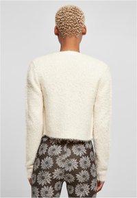 Krämfärgad, fluffig kort tröja med långa ärmar, texturerad yta och öppen ryggdesign, kombinerad med bruna byxor med blommönster.