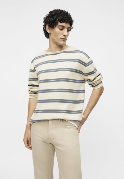 Jeune homme aux cheveux bouclés portant une chemise à manches longues rayée crème et grise et un pantalon beige, regardant vers le bas avec la main dans la poche.