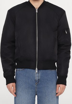 Person trägt eine schwarze Bomberjacke mit einem silbernen Frontreißverschluss und blauen Jeans, steht frontal.