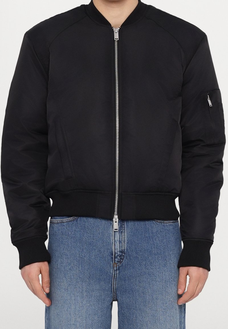 Personne portant une veste bomber noire avec une fermeture éclair argentée à l'avant et un jean bleu en denim, debout face à l'avant.