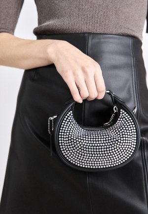 Ruka držící malou černou kabelku ve tvaru půlměsíce se stříbrnými kulatými nýty, oblečená v černé kožené sukni a hnědém žebrovaném topu.