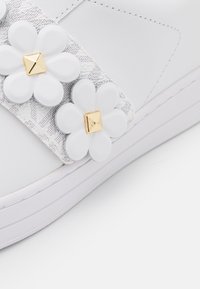 Détail d'une basket blanche avec une bande grise texturée et deux ornements en forme de fleurs blanches avec des clous pyramides dorés sur le bord de la semelle.
