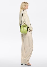 Mała zielona skórzana torebka z uchwytem u góry i regulowanym paskiem. Charakteryzuje się błyszczącą teksturą i nadrukowanym logo. Model nosi jasnobeżowe, luźne spodnie.