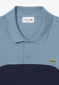 Two-tone polo shirt with light blue top, navy bottom, button placket, and green embroidered Lacoste crocodile logo on chest.