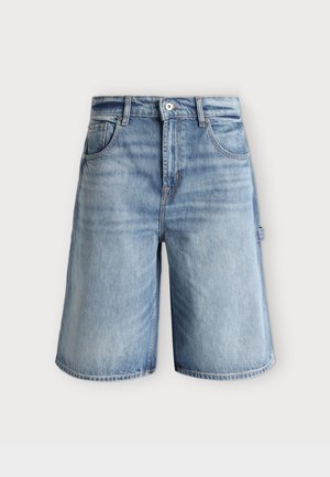 Jeune homme portant un sweat-shirt gris clair à demi-fermeture éclair, un short en jean longueur genou, des chaussettes blanches et des mocassins en cuir noir sur fond blanc uni.