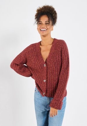 Cardigan rouge à côtes avec un décolleté en V profond, de gros boutons et des manches larges. Porté sur un jean bleu clair, le tissu texturé offre de la chaleur.