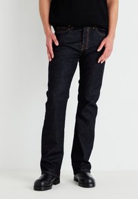 Jeans de ganga azul-escuro com um corte direito, costura visível e rebites de cor cobre. Usados com uma camisola preta com nervuras e botas pretas.