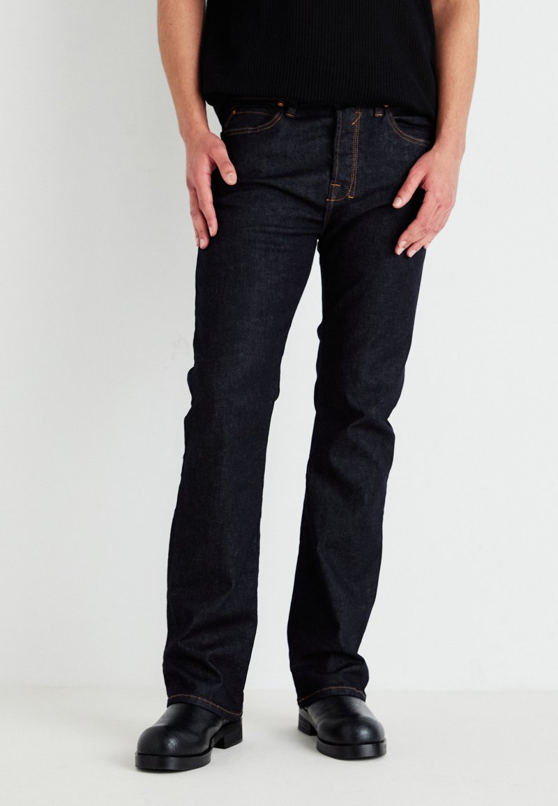 Jeans de ganga azul-escuro com um corte direito, costura visível e rebites de cor cobre. Usados com uma camisola preta com nervuras e botas pretas.