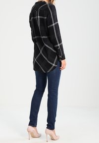Camicia a quadri nera e grigia con maniche lunghe, orlo alto-basso e spacchi laterali, abbinata a jeans blu scuro e tacchi nudi.