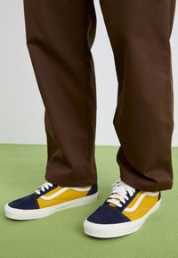 Blue and yellow sneakers with suede and canvas materials, featuring white laces, a white stripe design, and a rubber sole. Brown pants are visible.