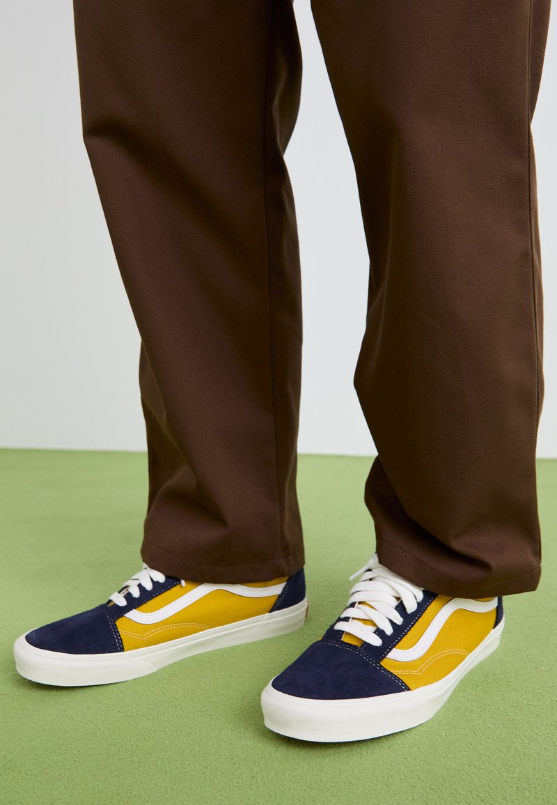 Blue and yellow sneakers with suede and canvas materials, featuring white laces, a white stripe design, and a rubber sole. Brown pants are visible.