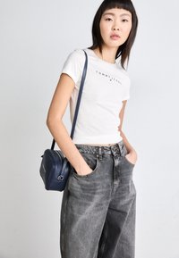 Bílý zkrácený tričko s logem "TOMMY JEANS", v kombinaci s vysokými šedými širokými džíny a malou námořnickou crossbody kabelkou.