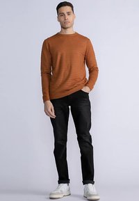Orangefarbenes, geripptes Langarmshirt, schwarze, eng anliegende Jeans und weiße Turnschuhe. Das Modell steht aufrecht mit den Händen in den Taschen vor einem hellen Hintergrund.