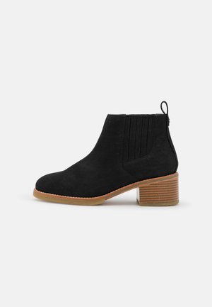 Bottine en suede noir avec côtés en élastique nervuré, bout arrondi et talon en bois empilé. Équipée d'une languette pour un enfilage facile.